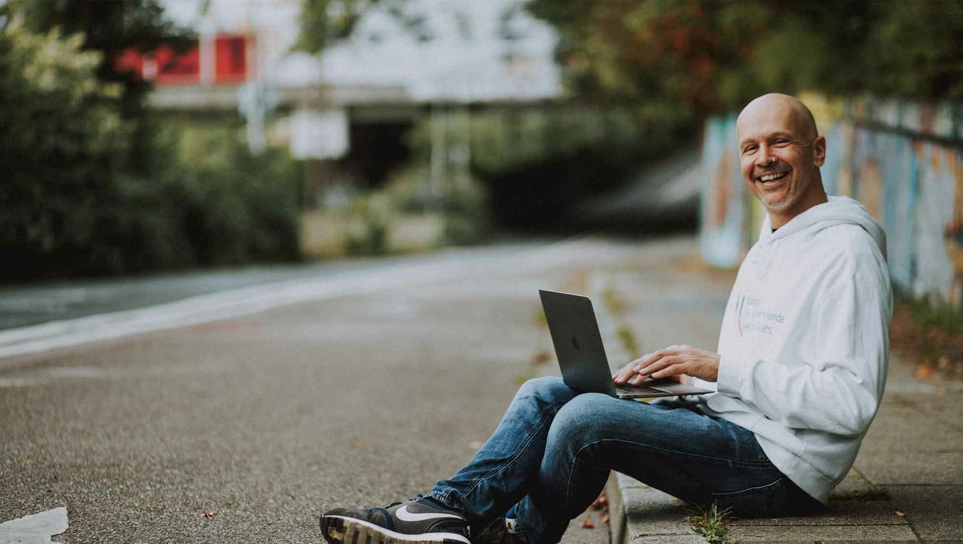 Web designer and graphic designer Paul Jackson sits on the kerb with his laptop and smiles